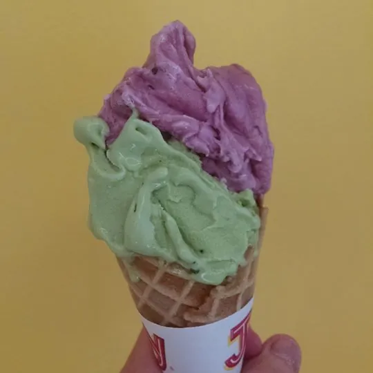 A hand holding a cone with a scoop of purple grape on green tea gelato