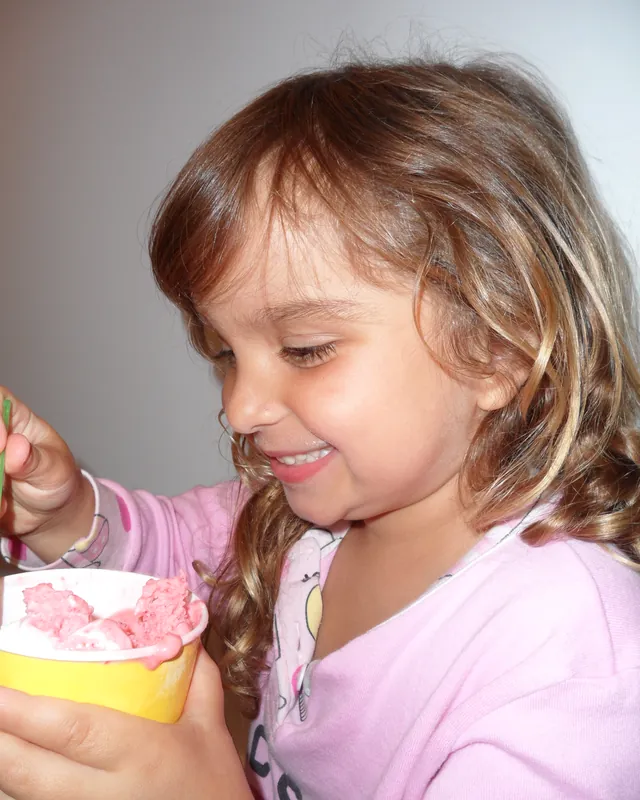A little girl smiling down at a yellow cup of pink gelato, spoon in hand